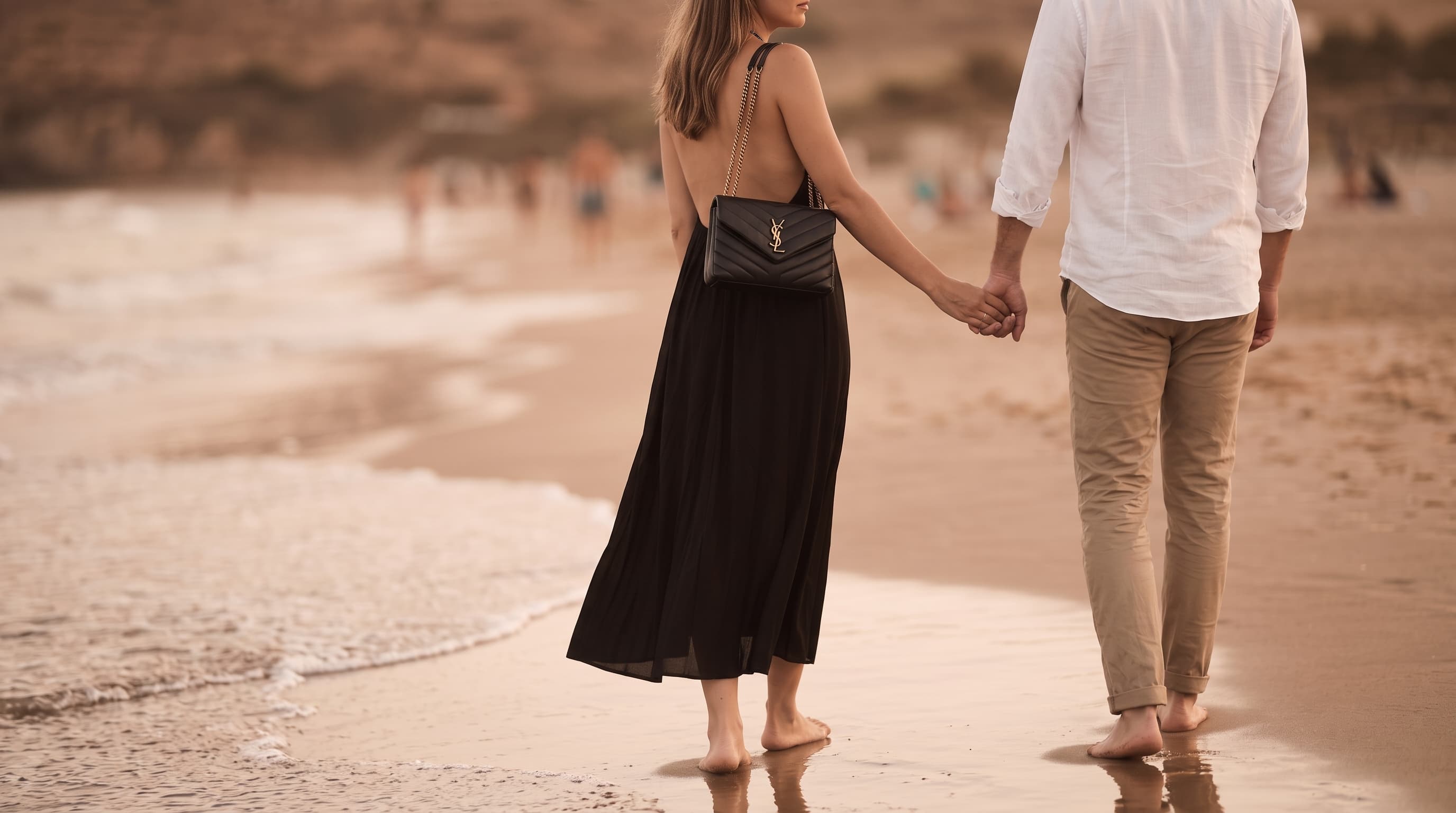 A couple walking hand in hand along a beach at golden hour, the woman carrying a designer purse