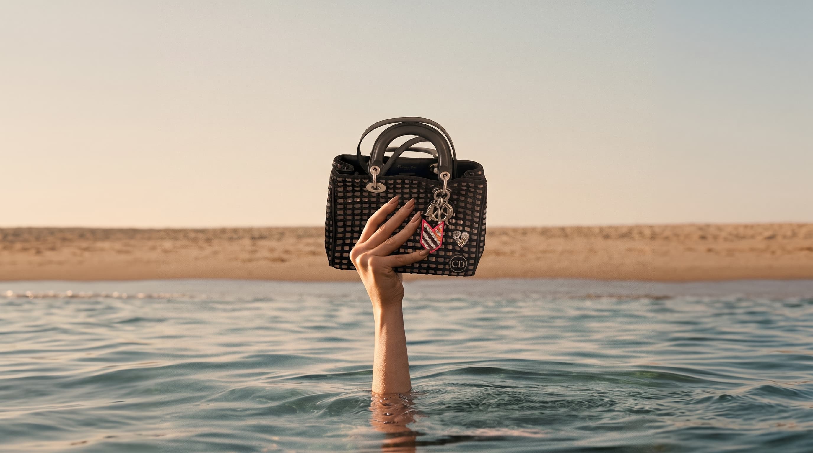A hand emerging from calm water holding a vintage Dior tote, sandy shore and pale sky above